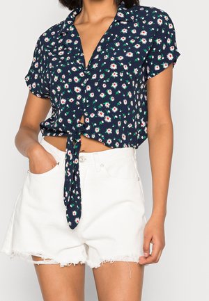 Woman wearing a navy blue short-sleeve cropped shirt with white daisy print tied at the waist and white frayed denim shorts.