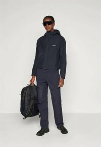 Žena v čiernych slnečných okuliaroch, čiernej kapucni, čiernych nohaviciach a čiernych topánkach, drží veľký čierny batoh pri svojom boku.