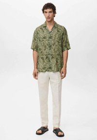 Camisa verde de manga corta con un patrón geométrico de hojas, combinada con pantalones crema y sandalias negras, creando un look casual y relajado.