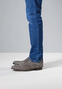 Harmaat ja mustat kuvioidut kengät, joissa on matala korko ja pyöreä kärki, yhdistetty sinisiin farkkuihin. Pinta on teksturoitu, kengännauhat solmittuina.