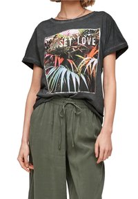 Femme portant un t-shirt gris foncé aux manches retroussées avec un imprimé tropical coloré et le texte "SUNSET LOVE", associé à un pantalon à cordon de serrage vert olive.