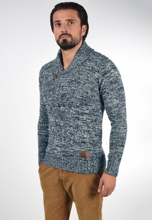 Homme aux cheveux foncés et à la barbe portant un pull en maille bleu-gris avec un col châle boutonné et un pantalon en velours côtelé marron.