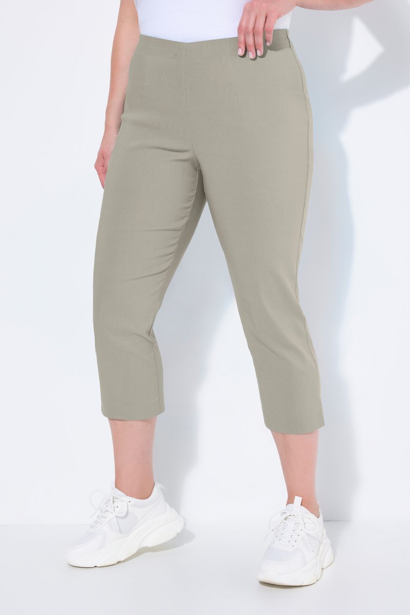 Frau trägt beige verkürzte Hose und weiße Sneaker und steht vor weißem Hintergrund mit einer Hand auf der Hüfte.