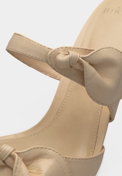 Κοντινή λήψη μπεζ δερμάτινου σανδάλι με λεπτομέρεια κόμπου στο λουράκι και ρυθμιζόμενο λουράκι αστραγάλου.