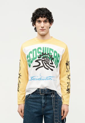 Νέος άνδρας με σγουρά μαλλιά που φοράει μακρυμάνικη μπλούζα GCDSwear κίτρινη και λευκή με μαύρα σχέδια ήλιου στα μανίκια και σκούρα μπλε τζιν.