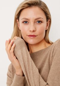 Maglione in cotone leggero, di colore marrone chiaro, con polsini e scollatura a coste. La texture sembra morbida, con una vestibilità comoda intorno alle spalle e alle braccia.