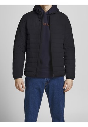 Homme portant une veste matelassée noire par-dessus un sweat à capuche bleu marine et un jean bleu, debout devant un fond gris clair uni.