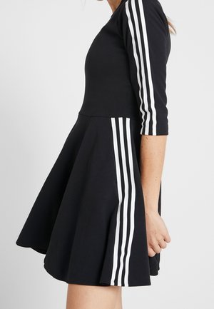 Femme portant une robe noire avec trois bandes blanches sur les manches et la jupe, debout devant un fond clair uni.