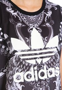 Svart Adidas-tröja med stort vitt Trefoil-logotyp och detaljerat mönster av gråskala på påfåglar och blommor i bakgrunden.