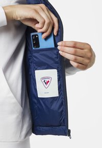 Mörkblå jackficka med en vit logotypklistermärke med röda och blå detaljer; en blå smartphone sätts in i fickan.