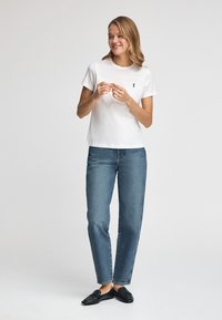 Vit bomullst-shirt med korta ärmar och en liten logga på vänster bröst, kombinerad med blå jeans med raka ben och svarta slip-on skor.