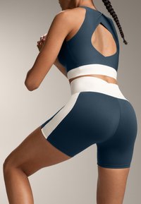 Námornícky modrý športový crop top s vyrezávaným zadným dizajnom, skombinovaný s vysokými šortkami v rovnakom odtieni s bielymi bočnými akcentmi. Hladká tkanina.