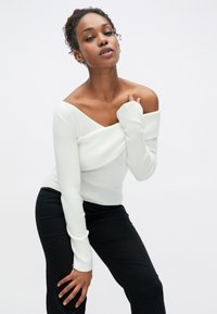 Pull-over blanc à épaules dénudées avec un détail torsadé, associé à un pantalon noir ajusté. Texture lisse ; manches longues et ajustées.