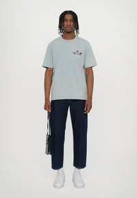 Gai zila īsām piedurknēm T-kreklu ar nelielu grafiku uz krūtīm, kombinēts ar tumšas denim biksēm un baltām sporta apaviem. Minimalistisks dizains.