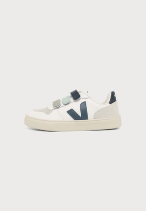 Zapatilla blanca con tres tiras de velcro de colores y un logo "V" en azul oscuro en el lateral, mostrada sobre un fondo claro y simple.