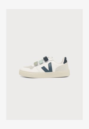 Zapatilla blanca con tres tiras de velcro de colores y un logo "V" en azul oscuro en el lateral, mostrada sobre un fondo claro y simple.