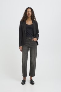 Kobieta stojąca, ubraną w czarną marynarkę, czarną bluzkę, wyblakłe czarne dżinsy i czarne mokasyny na jednolitym jasnym tle.