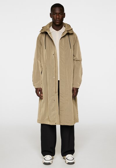 Beige, lätt lång kappa med huva, med en krinklad textur, framknappar, sidofickor och svarta dragsnören. Buren över en ljus skjorta och mörka byxor.