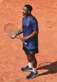 Camiseta atlética morada y negra con patrones ondulados, combinada con pantalones cortos negros. Sosteniendo una raqueta de tenis multicolor en una cancha de arcilla. Zapatillas negras.