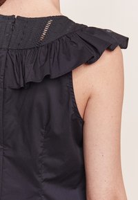 Černý top s volánkovým límcem, s texturovanou horní částí a hladkým, přiléhavým tělem. Zapínání na zip vzadu.