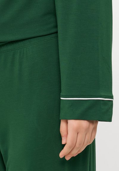 Green long-sleeve garment with a cuff featuring a white accent. The fabric appears soft with a smooth texture. Hand visible near the cuff.