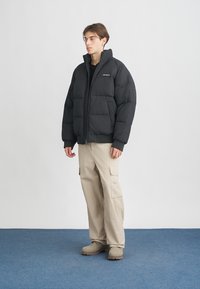 Fiatal férfi fekete lélegző dzsekiben, bézs cargo nadrágban és bézs csizmában áll egy sima világos fal előtt, kék szőnyeges padlón.