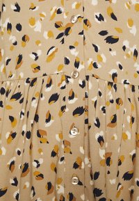 Tissu marron avec un motif texturé de feuilles noires, blanches et moutarde ; présente des boutons et des plis froncés à la taille.