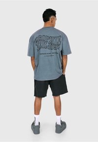 Gray oversized t-shirt with large black text on the back. Paired with dark shorts and gray sneakers, worn with white socks.