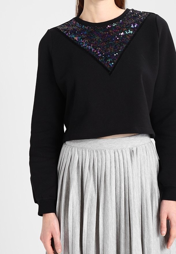Femme portant un top noir à manches longues avec un triangle pailleté scintillant et une jupe plissée taille haute gris clair.