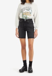 Grå sweatshirt med grafiskt tryck, högmidjade svarta denimshorts med fransad nederkant, och svarta träskor med ribbade ankelsockor.