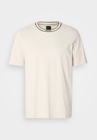 T-shirt crème à manches courtes avec un col rond, présentant un liseré noir contrastant le long du col et une étiquette à l'intérieur du col.