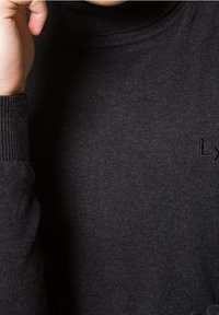 Pull à col roulé noir en tissu doux et texturé. Comprend un col et des poignets côtelés, petit logo brodé sur la poitrine.