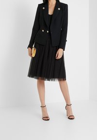 Blazer nero con accenti di bottoni dorati, abbinato a una gonna di tulle nera e tacchi neri con cinturini. Tiene una piccola pochette dorata.