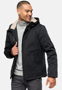Chaqueta negra con capucha, con un cuello de borreguillo beige, dos grandes bolsillos frontales y un cierre de cremallera con costuras contrastantes.