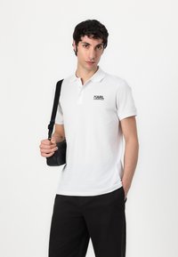 Valkoinen polo-paita lyhyillä hihoilla ja kauluksella, jossa on musta "KARL LAGERFELD" -logo. Musta laukku säädettävällä hihnalla kädessä.