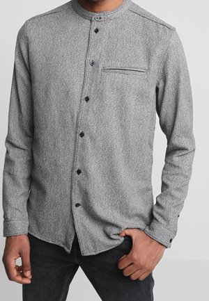 Mann trägt ein hellgraues, strukturiertes Hemd mit Knöpfen und Stehkragen sowie schwarze Hose, Hand justiert den Hemdsaum.