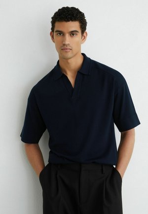 Giovane uomo con capelli ricci che indossa una polo blu scuro a maniche corte e pantaloni neri, in piedi con le mani in tasca contro un muro bianco.
