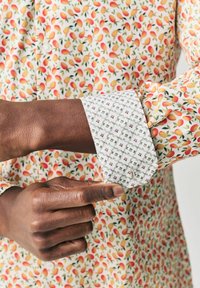 Camisa de cuello con un patrón afrutado en rojo y amarillo sobre una base blanca. El puño es blanco con un estampado geométrico en tonos gris y burdeos.
