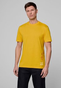 Żółta bawełniana koszulka z okrągłym dekoltem, krótkimi rękawami oraz czerwonym nadrukiem z boku i logo przy krawędzi.