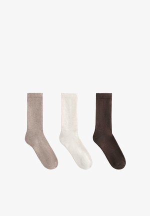 Trois chaussettes à côtes de couleur beige, blanche et brun foncé, présentées côte à côte sur un fond blanc.