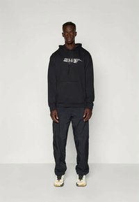 Černá mikina s kapucí a šňůrkou, na hrudi s grafikou, v kombinaci s černými kalhotami. Oblečení doplňují bílé a žluté tenisky.