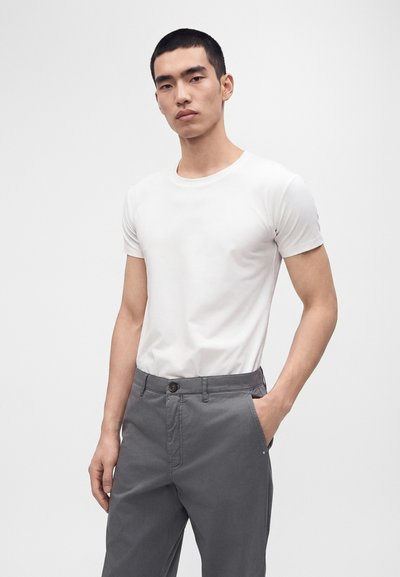 Jeune homme portant un t-shirt blanc ajusté à manches courtes et un pantalon gris, debout avec une main dans la poche devant un fond blanc uni.