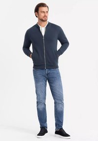 Felpa con zip di un blu scuro, con colletto a costine, tasche e una texture liscia, abbinata a jeans di denim chiaro e sneakers nere.