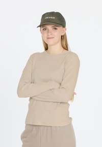 Beige gestreiftes Langarmshirt, kombiniert mit passenden Jogginghosen, akzentuiert durch eine dunkelgrüne Baseballmütze mit dem Stickmuster "LES'ARG" auf der Vorderseite.