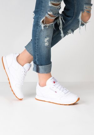 Gambe con jeans blu strappati arrotolati e sneakers bianche Reebok con suole color gomma, posizionate contro uno sfondo chiaro e uniforme.
