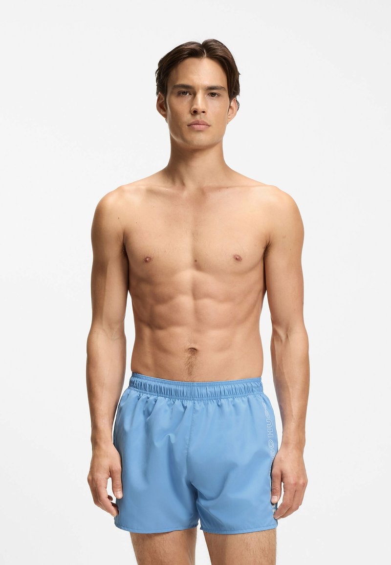 Jeune homme aux cheveux courts bruns portant un short de bain bleu clair, debout devant un fond blanc.