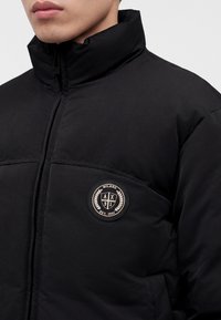 Personne portant une doudoune noire avec un col haut et un patch rond du logo Milano A|X 91 sur la poitrine.