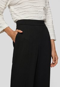 Schwarze Hose mit weitem Bein aus strukturiertem Stoff, hohem Bund und Seitentaschen, kombiniert mit einem weißen, gerippten Langarmoberteil.