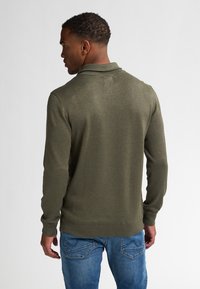 Olijfgroene polo met lange mouwen, fijne brei structuur, geribde kraag en manchetten, gedragen met blauwe denim jeans.