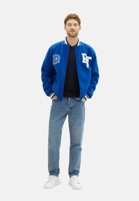 Blaues Varsity-Jackett mit weißen Akzenten, Logodetail und Druckknöpfen; kombiniert mit hellen Jeans und weißen Sneakers. Einfaches Design.